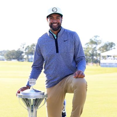 Tony Finau is kneeling down and has one hand on the trophy.